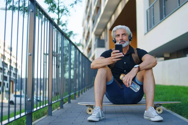 Portret dojrzałego mężczyzny ze słuchawkami siedzącymi na świeżym powietrzu w mieście, za pomocą smartfona. — Zdjęcie stockowe