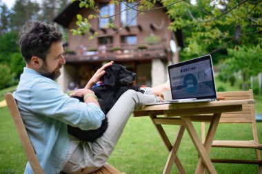 Mature man with laptop and dog working outdoors in garden, home office concept. clipart