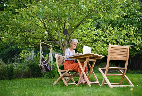 Starsza kobieta z laptopem pracująca przy stole w ogrodzie, koncepcja biura domowego. — Zdjęcie stockowe
