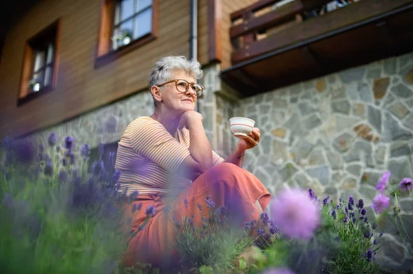Portret van een oudere vrouw die in de tuin zit en een kop koffie vasthoudt. — Stockfoto