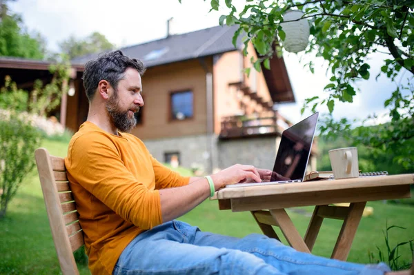 Sidovy av man med bärbar dator arbetar utomhus i trädgården, hemmakontor koncept. — Stockfoto