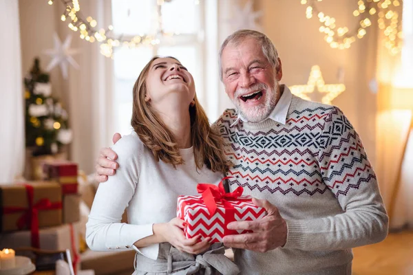 Homem sênior com jovem mulher em casa em casa no Natal, segurando presente. — Fotografia de Stock