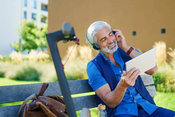 Porträt eines reifen Mannes mit Kopfhörern, der draußen in der Stadt sitzt und ein Tablet benutzt. — Stockfoto