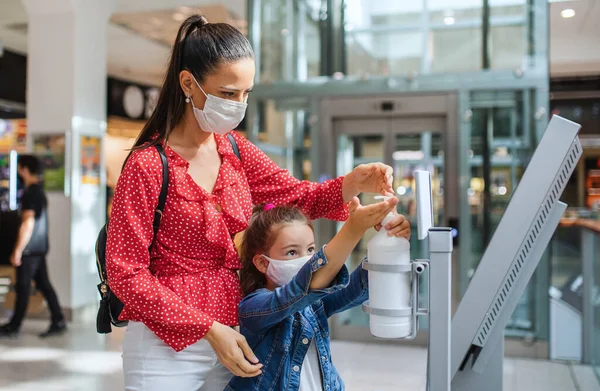 Mother and daughter with face mask disinfecting hands indoors in shopping center, coronavirus concept. — Stock Photo, Image