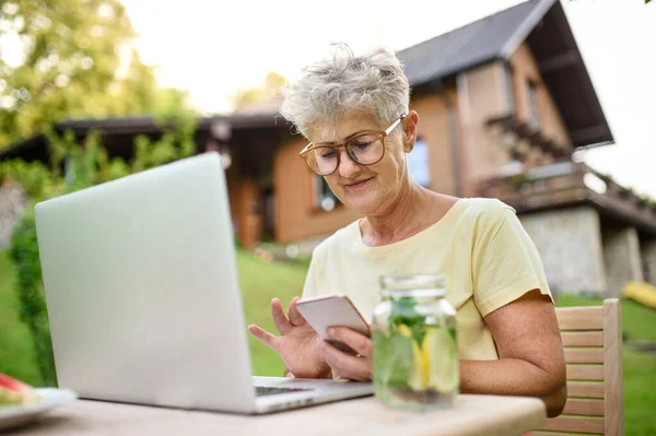 Пожилая женщина с ноутбуком и смартфоном, работающая в саду, концепция домашнего офиса. — стоковое фото