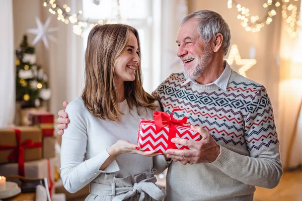 Homme âgé avec jeune femme à l'intérieur à la maison à Noël, tenant présent. — Photo