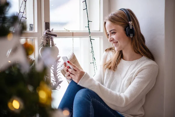 Porträt einer jungen Frau zu Weihnachten zu Hause mit Smartphone und Kopfhörer. — Stockfoto