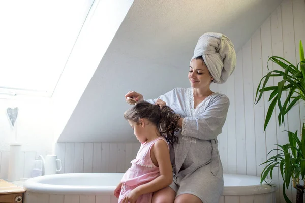 Portrait of pregnant woman with small daughter indoors in bathroom at home, brushing hair. — Stock Photo, Image