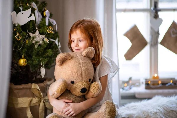Mała dziewczynka stojąca w domu w święta, przytulająca pluszowego misia. — Zdjęcie stockowe