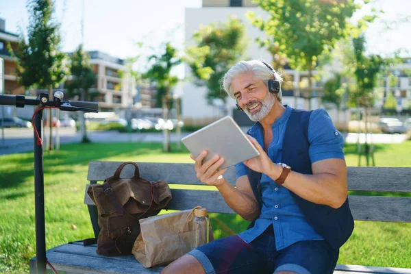 Портрет дорослого чоловіка з навушниками, що сидять на відкритому повітрі в місті, використовуючи планшет . — стокове фото