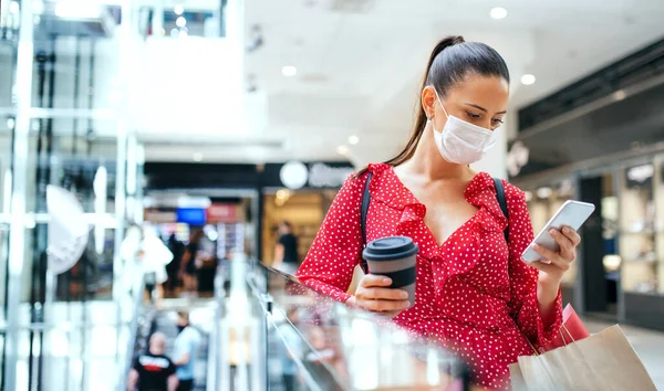 Woman with face mask standing and using smartphone indoors in shopping center, coronavirus concept. — Stock Photo, Image