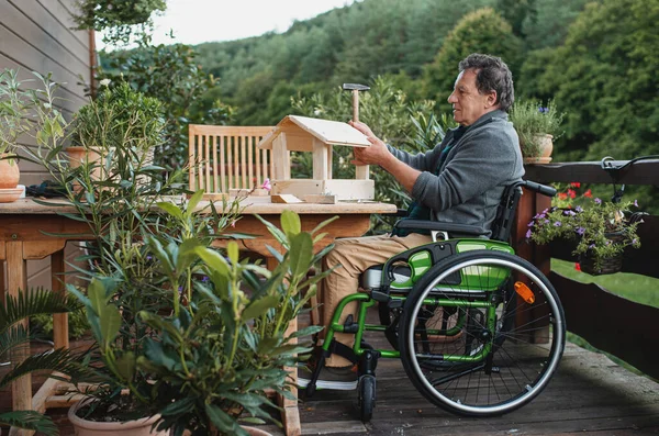Старший в инвалидной коляске строит скворечник на открытом воздухе на террасе, diy project. — стоковое фото