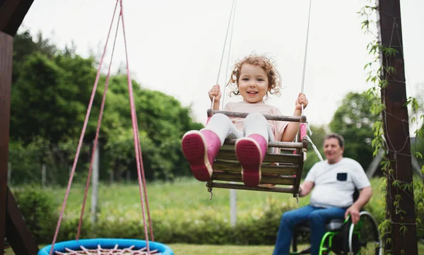 Маленька дівчинка з дідусем у інвалідному візку грає у саду на задньому дворі, гойдається . — стокове фото