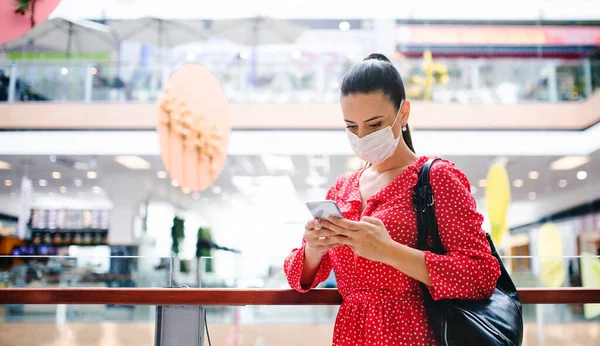 Žena s obličejovou maskou stojící s chytrým telefonem uvnitř v nákupním centru, koncepce koronaviru. — Stock fotografie