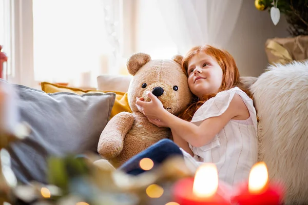 Menina pequena sentada no sofá dentro de casa no Natal, abraçando ursinho de pelúcia. — Fotografia de Stock