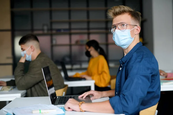 Jovens estudantes com máscaras faciais em mesas na faculdade ou universidade, conceito coronavírus. — Fotografia de Stock