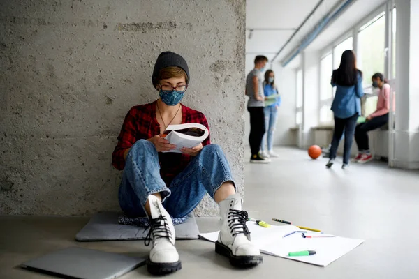Молодой студент в маске для лица сидит на полу в колледже или университете, коронавирусная концепция. — стоковое фото