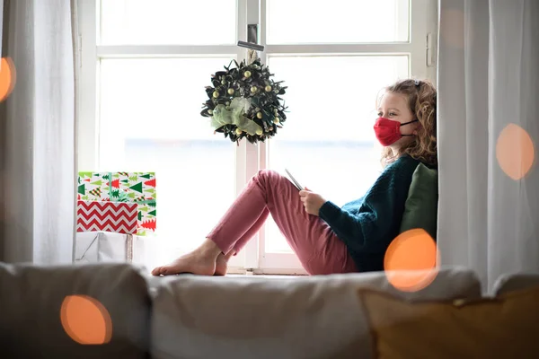 Kleines Mädchen mit Gesichtsmaske sitzt zu Weihnachten zu Hause und bedient sich eines Tablets. Coronavirus-Konzept. — Stockfoto