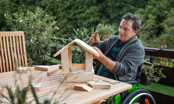 Starszy mężczyzna na wózku inwalidzkim budujący domek dla ptaków na tarasie, projekt. — Zdjęcie stockowe