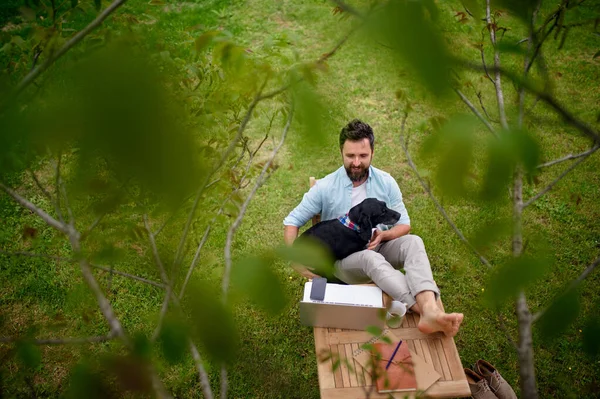 Uomo d'affari con cane e laptop che lavora all'aperto in giardino, concetto di home office. — Foto Stock