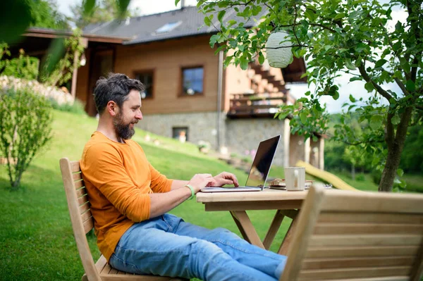 Widok z boku człowieka z laptopem pracy na zewnątrz w ogrodzie, koncepcja biura domu. — Zdjęcie stockowe