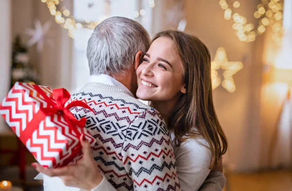 Ung kvinna ger present till glad farfar inomhus vid jul. — Stockfoto