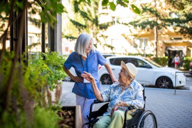 Tekerlekli sandalyedeki yaşlı kadın, bakıcı asistanıyla temiz havanın tadını çıkarıyor..