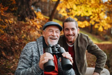 Kıdemli baba ve oğlu dürbünle doğada bankta otururken, konuşuyor.