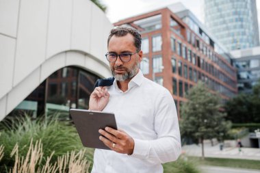Modern ofis binasının önünde elinde tablet olan ve bir şeyler okuyan bir iş adamı..