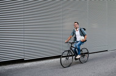 Sokakta bisiklet süren, sırt çantası takan bir banliyö. Kameraya bakıp gülümsüyor..