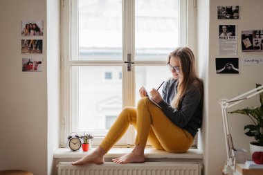 Ders kitabı okuyan kız öğrenci pencere eşiğinde oturup ders çalışıyor..