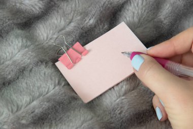 Mavi manikür ile genç kız pembe kalem tutun ve gümüş gri tüylü faux kürk battaniye arka plan üzerinde pembe metal kelepçe Kırtasiye ile hafif pembe etiket üzerine yazmak.