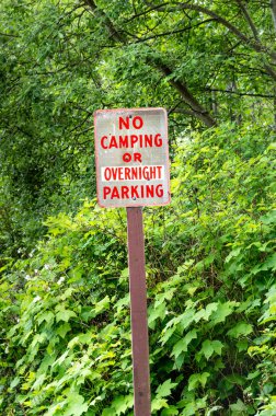 Yeşil ağaçların yanında kırsal yolda kırmızı harflerle kamp veya geceleme park tabelası yok.