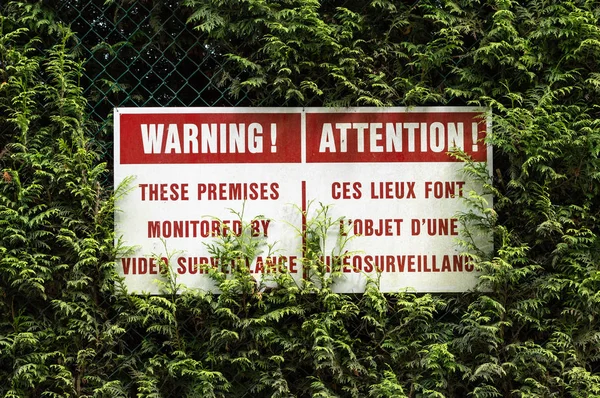 English and French surveillance warning sign with red lettering hung on chain link fence in green cedar hedge.