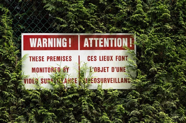 English and French surveillance warning sign with red lettering hung on chain link fence in green cedar hedge.