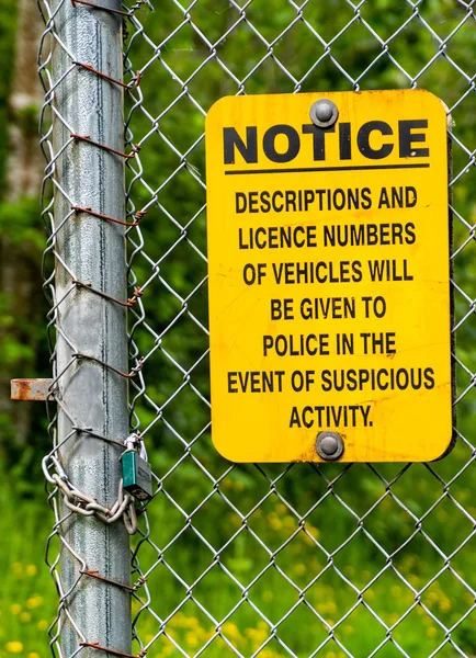 Yellow and black neighborhood crime warning sign posted on chain link fence with wooded background.