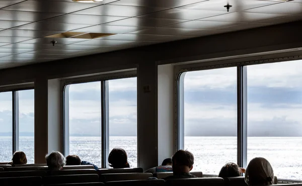 Silhouettes of indoor boat passengers seated beside bright oceanview window.