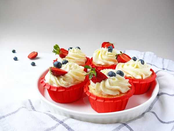 Beyaz doku arka planda ve modamutfak havlusunda krem peynir, çilek ve yaban mersini ile cupcakes.