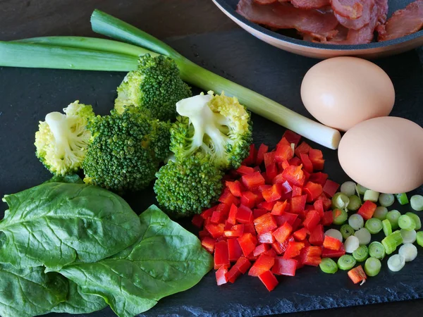 Verduras cortadas en cubitos brócoli, espinacas, pimiento rojo, tocino y huevos, ingredientes para tortilla en pizarra negra . — Foto de Stock