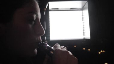 Gece vakti şehrin aydınlanmış arka planında elektronik sigara içen bir kadın.