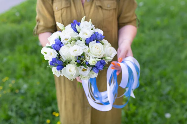Delicate bouquet of fresh flowers with ribbons in the hands of a girl on a background of grass (Eustoma. Primary colors: white, blue)