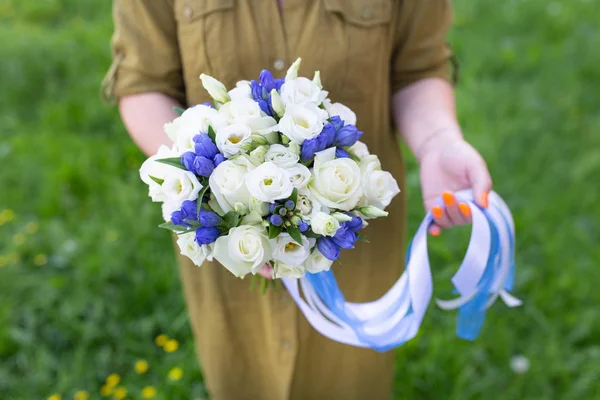 Delicate bouquet of fresh flowers with ribbons in the hands of a girl on a background of grass (Eustoma. Primary colors: white, blue)