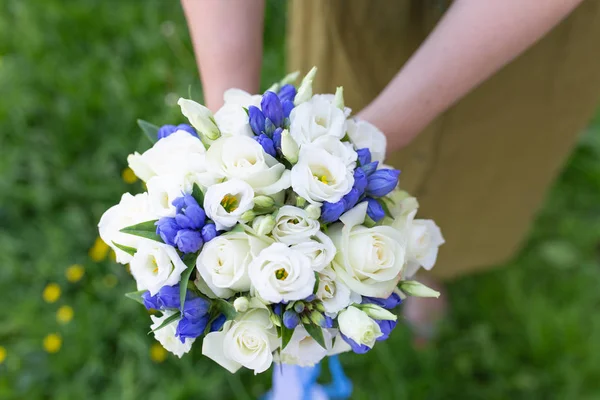 Delicate bouquet of fresh flowers with ribbons in the hands of a girl on a background of grass (Eustoma. Primary colors: white, blue)