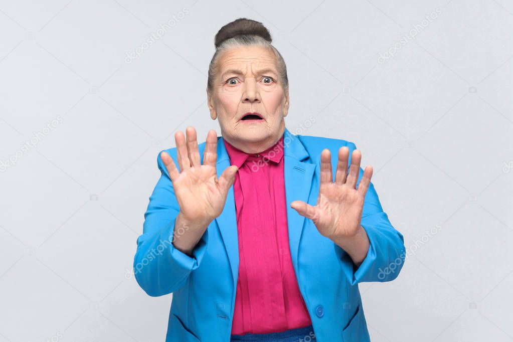 scared aged woman with collected hair in blue suit and pink shirt looking at camera with shocked face and big eyes on grey background