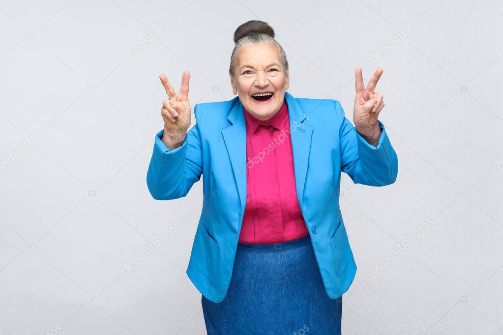 Funny aged woman wearing blue suit and pink shirt laughing and showing peace signs at camera on gray background