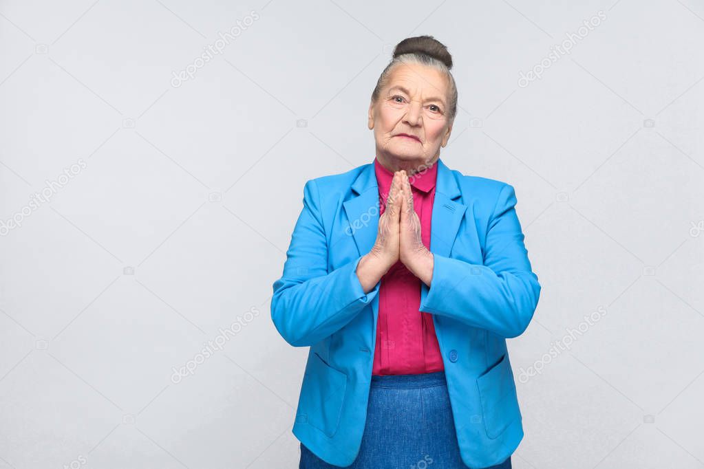 expressive grandmother wearing blue suit and pink shirt holding hands in apologize gesture on gray background