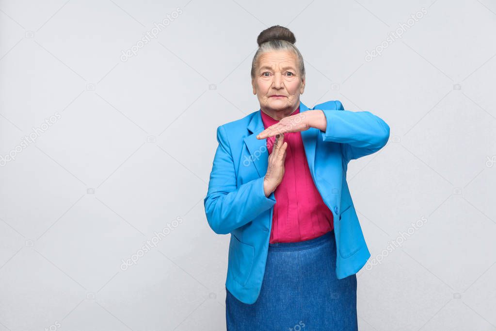 expressive grandmother with bun hairstyle in blue suit and pink shirt showing time out sign on gray background 