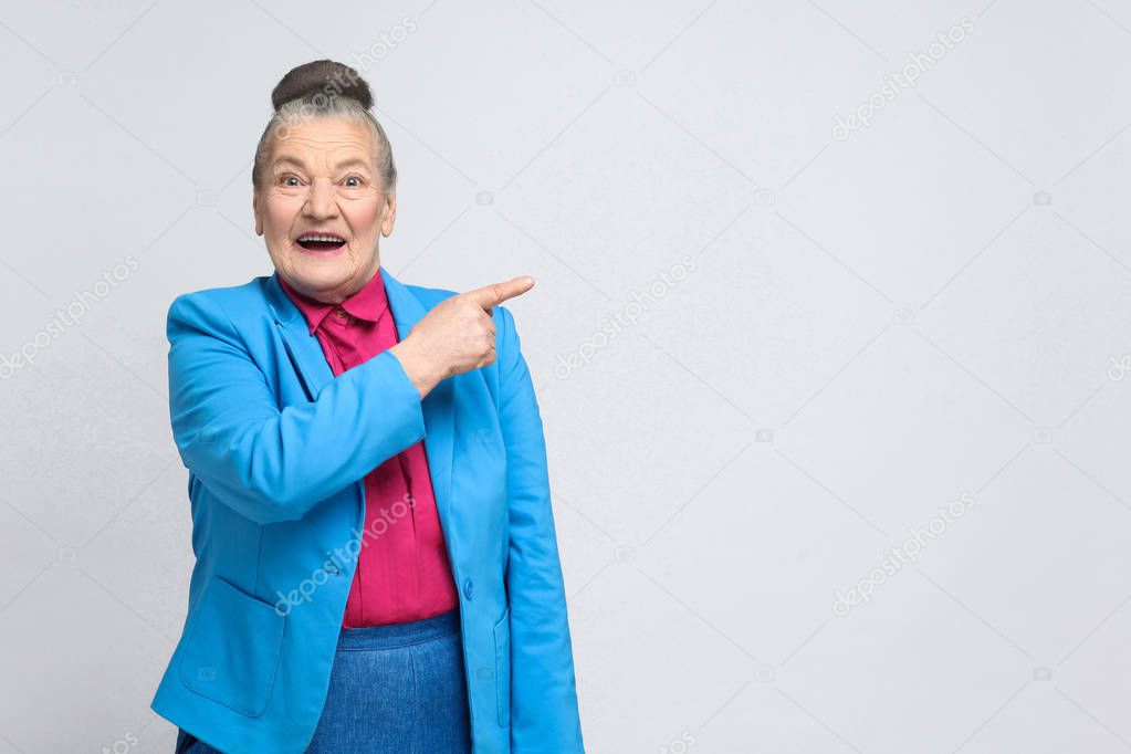 aged woman in blue suit and pink shirt pointing finger to right and toothy smiling, Emotion and feelings concept