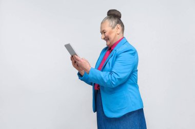 Profil görünümü tablet holding ve memnun yüz ve dişlek gri arka plan üzerinde gülümseyen ile ekrana bakarak yaşlı kadın