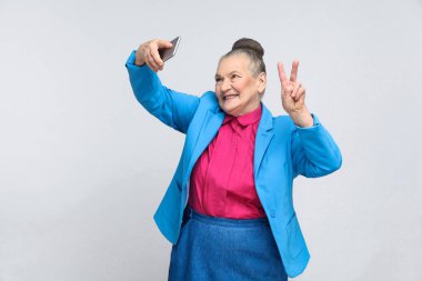 şarkı selfie ile barış yapma ve gri arka plan üzerinde gülümseyen toplanan gri saç topuz saç modeli ile ışık mavi elbiseli ifade Büyükanne